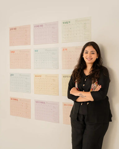 A woman with long dark hair stands with arms crossed, wearing a black blazer and trousers, standing in front of a white wall decorated with pastel-colored monthly calendar sheets. 