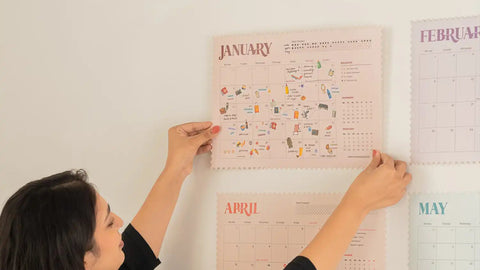 A woman in a black long-sleeve top hangs a pastel pink monthly calendar poster on a white wall. Other monthly calendar posters in soft pastel colors are partially visible around her.