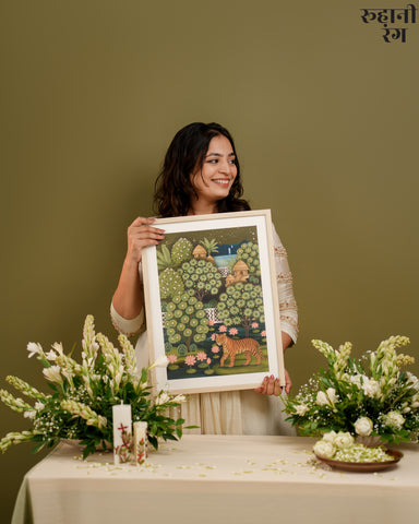 Woman holding a framed artwork with a green and brown color scheme, surrounded by flowers on a table.