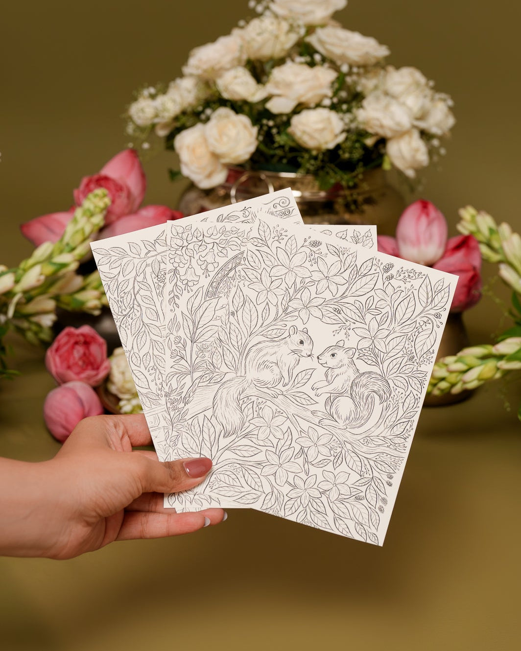 Hand holding two white cards with black patterns in front of floral arrangements on a brown background