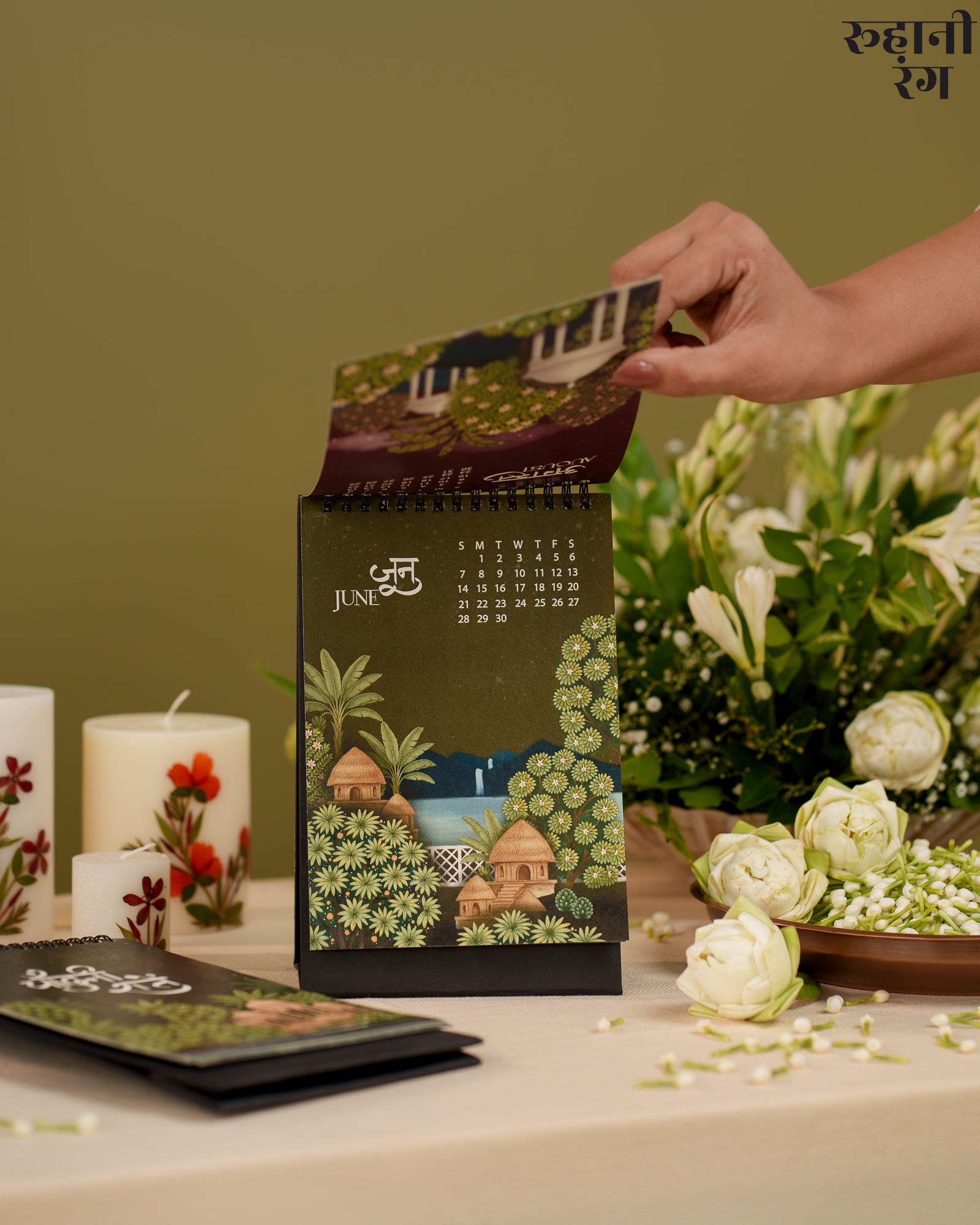Hand opening a decorative calendar with floral and architectural design, candles, and flowers in the background.