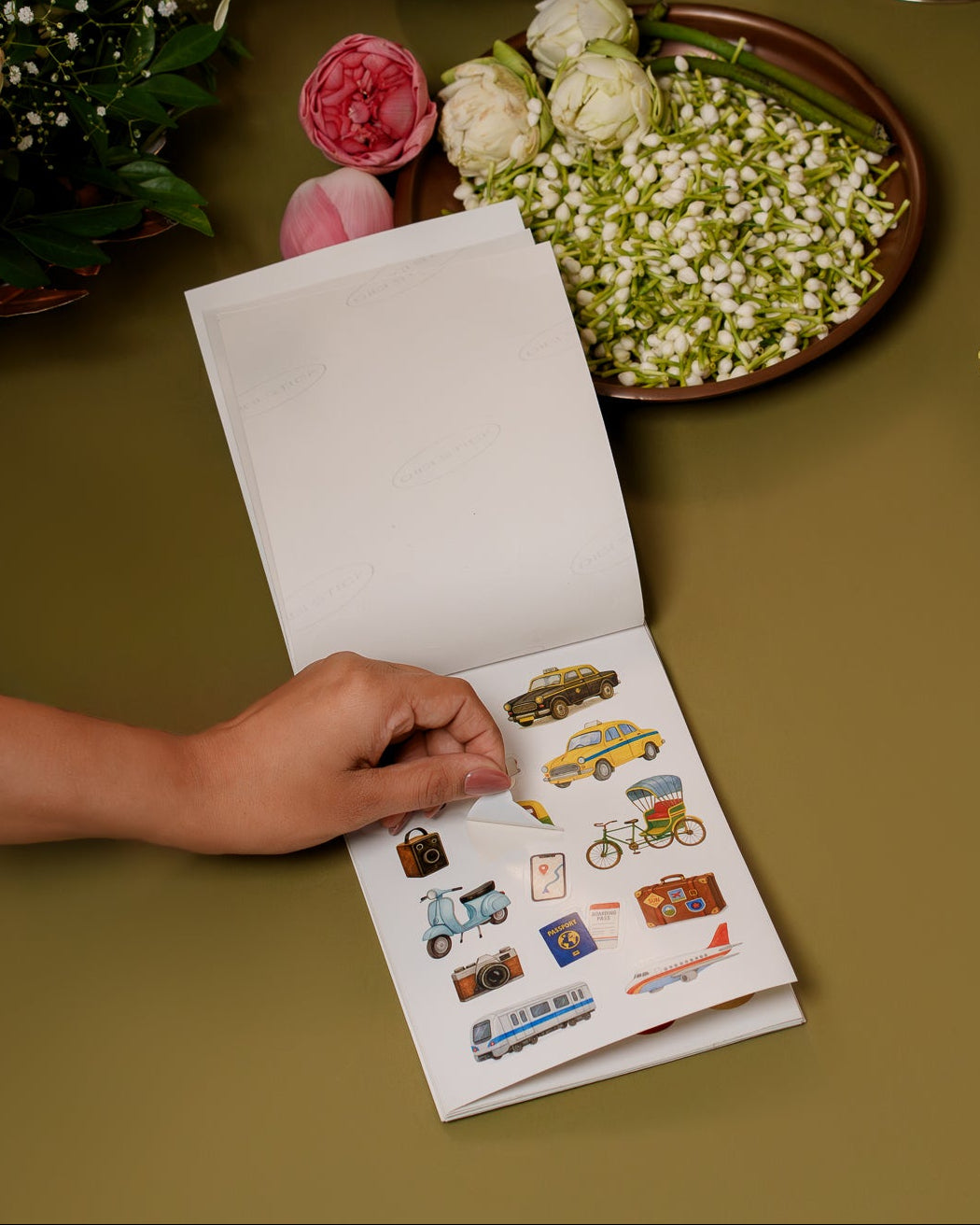 Person holding a small album with stickers on a table surrounded by flowers