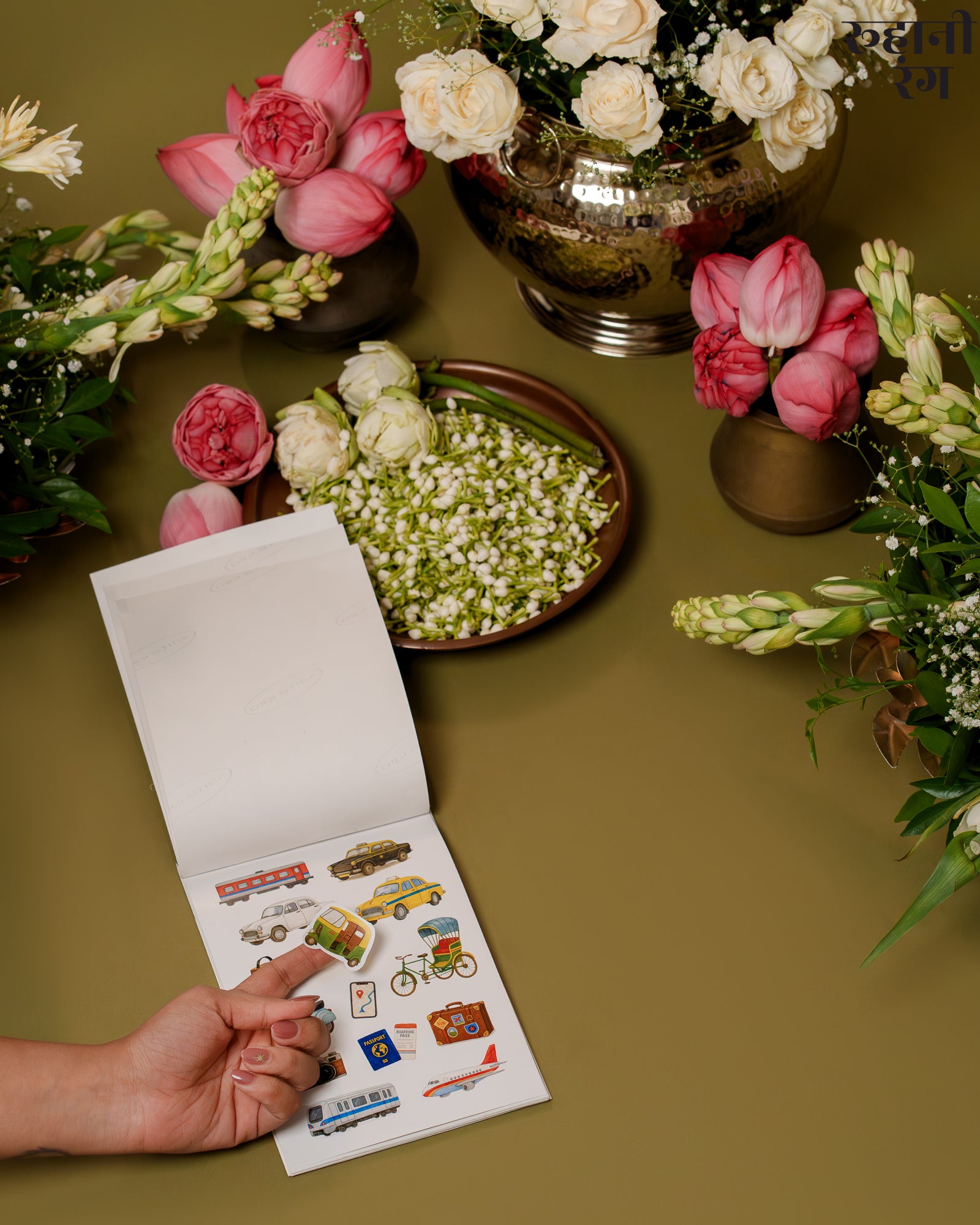 Hand holding a small open book with stickers on a table surrounded by flowers
