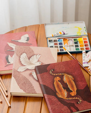 2027 planner and calendar in india . Three illustrated sketchbooks with bird and leopard artwork arranged on a wooden table beside an open watercolor palette, paint tubes, and brushes.